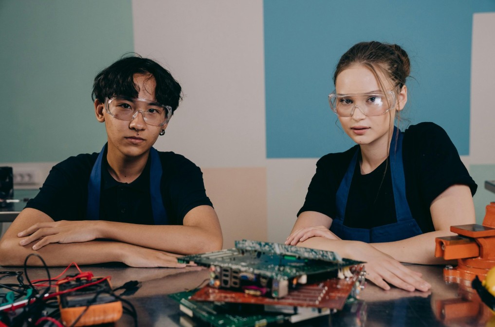 Zwei Jugendliche mit Schutzbrillen und Arbeitsschürzen sitzen an einem Labortisch mit elektronischen Platinen und Werkzeugen. Sie interessieren sich für MINT und Technik
